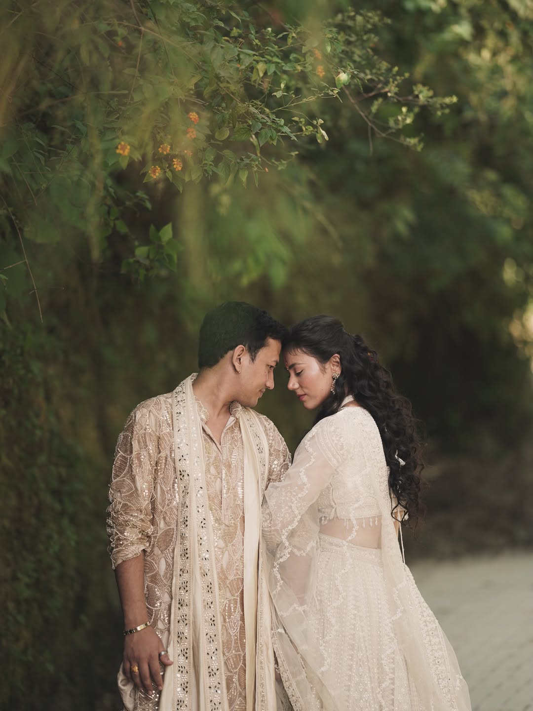 An engaged couple in ivory sharing a quiet moment beneath the trees, photographed by Nitin Bisht